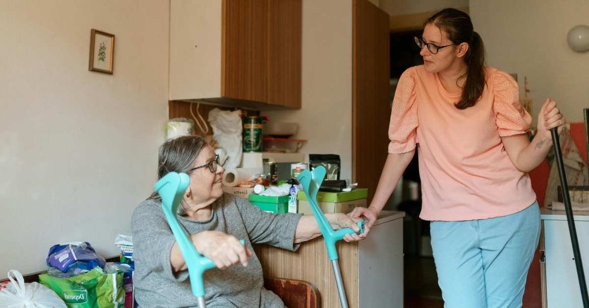 A caregiver helping an elderly woman in a cluttered home while providing light housekeeping services to support safe and comfortable living at home.