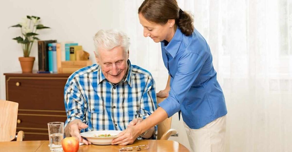 Paid family caregiver assisting an elderly man with meal support at home while balancing work and caregiving responsibilities.