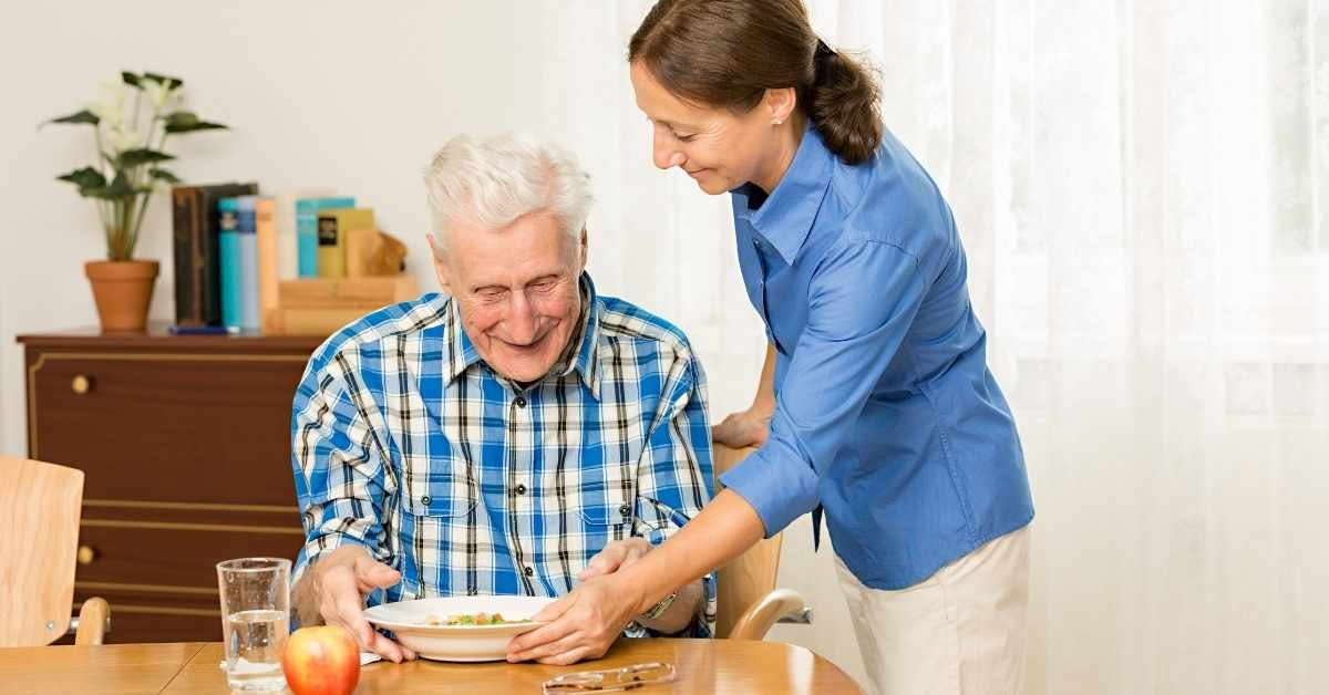 Paid family caregiver assisting an elderly man with meal support at home while balancing work and caregiving responsibilities.